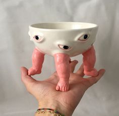 a hand holding a small white bowl with eyes on it's face and legs