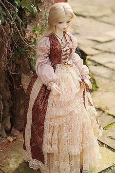 a doll dressed in an old fashion dress standing next to a tree and brick wall