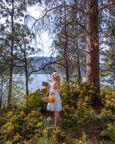 a woman in a white dress is standing by some trees and flowers near the water