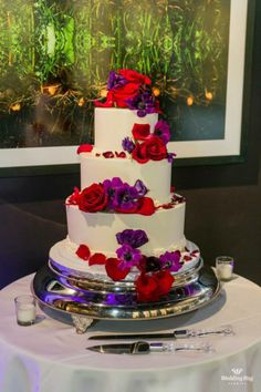 a three tiered wedding cake with red and purple flowers on the top is sitting on a table