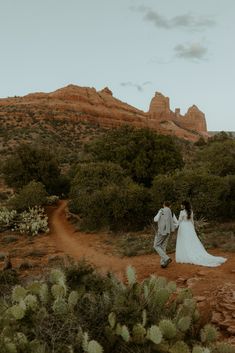 a bride and groom walking down a dirt path in front of the desert with mountains behind them