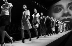 a group of women walking down a runway in front of a large screen with a woman's face on it