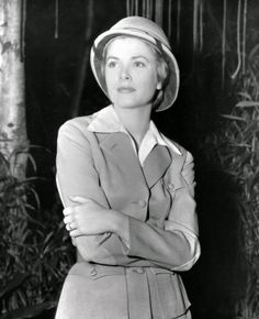 a woman wearing a hat and coat standing in front of some bamboo trees with her arms crossed