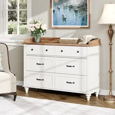 a white dresser with flowers and books on it next to a lamp in a living room