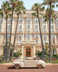 an old white car parked in front of a large building with palm trees around it