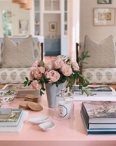 a table topped with books and flowers on top of a pink table cloth next to two couches