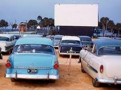 several old cars parked in a parking lot with a movie screen on the wall behind them