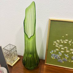 a green glass vase sitting on top of a wooden table next to a framed painting