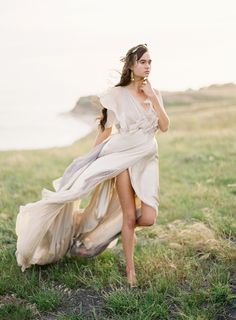 a woman in a white dress is walking on the grass by the ocean with her hair blowing in the wind