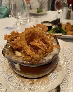 a hamburger with onion rings on it sitting on a plate next to wine glasses and utensils