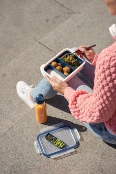 a woman sitting on the ground holding a tray with food in it and two containers next to her