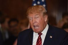 a man in a suit and tie making a face while speaking into a microphone with an american flag behind him