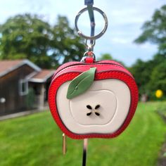 an apple shaped keychain hanging from a hook in the grass with a house in the background