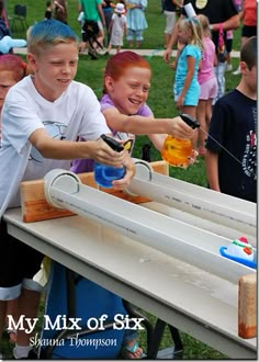 two young boys are playing with toys on a table at an outdoor event while others watch