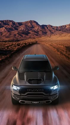 the front end of a black truck driving down a dirt road with mountains in the background