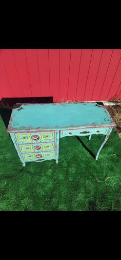 an old desk painted blue and yellow in front of a red building with grass on the ground