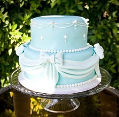 a three layer blue cake with pearls and bows on a glass platter in front of some bushes