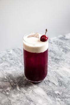 a purple drink with a cherry on the top sits on a marble countertop in front of a white wall