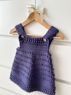 a purple crocheted dress hanging on a white wall with a wooden hanger