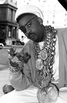 a black and white photo of a man with jewelry on his neck sitting at a table