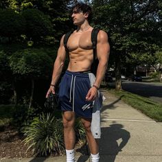 a man with no shirt and shorts standing on the sidewalk in front of some trees