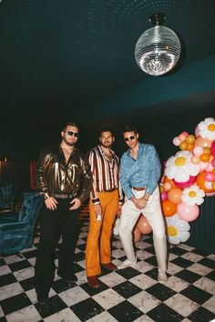 three men standing next to each other in a room with balloons on the wall and a disco ball hanging from the ceiling