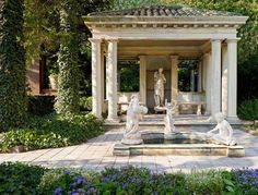 an outdoor fountain surrounded by greenery with statues in the center and blue flowers around it