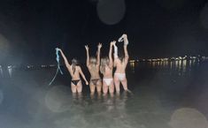 four women in bikinis are standing in the water at night with their arms up