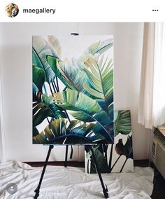 an easel sitting on top of a white bed next to a painting with green leaves