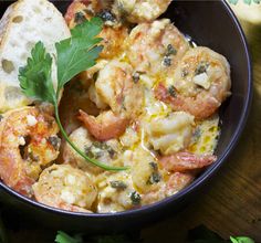 a bowl filled with shrimp and bread on top of a wooden table next to parsley