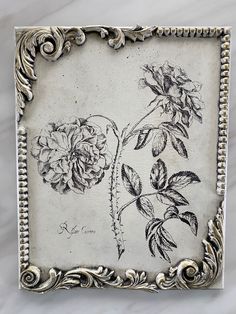 an ornate silver frame with flowers and leaves on the inside, in front of a marble background