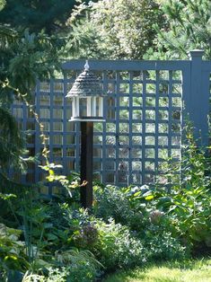 a bird feeder in the middle of a garden with plants and trees around it, next to a fence