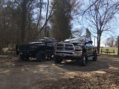 two large trucks parked next to each other on a dirt road in front of trees