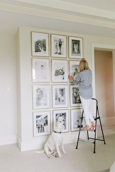 a woman standing on a ladder in front of a wall full of pictures and a dog