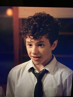 a young boy wearing a white shirt and black tie with curly hair smiling at the camera