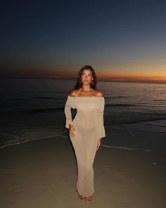 a woman standing on top of a sandy beach next to the ocean at sunset with her hands in her pockets