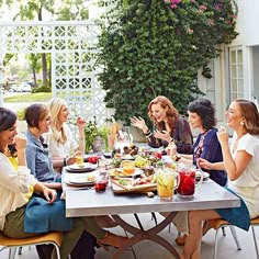 a group of women sitting around a table eating food and drinking wine, talking to each other