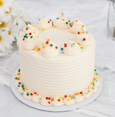 a white frosted cake with sprinkles sits on a plate next to flowers