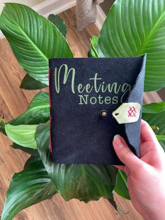 a hand holding a black notepad with writing on it and a green plant in the background