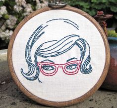 a hand embroidered hoop with a woman's face and glasses on it, sitting next to a potted plant