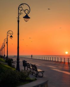 the sun is setting over the ocean with benches and street lamps in front of it