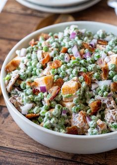 a bowl filled with peas and bacon on top of a wooden table next to plates