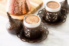 two cups of hot chocolate sit on saucers next to a copper pitcher and wooden cutting board