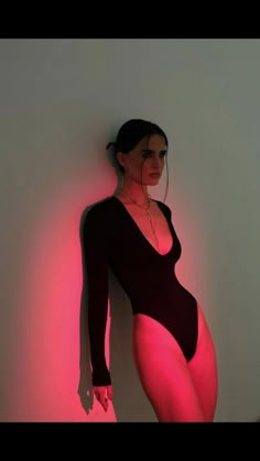 a woman in a black bodysuit standing next to a wall with pink light on it