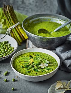 two bowls filled with green pea soup next to asparagus and other vegetables on a table