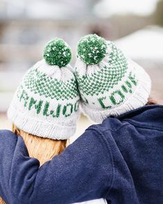 two knitted hats on the back of a person's head, both with green and white pom - poms