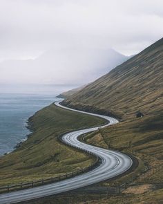 a winding road on the side of a grassy hill next to the ocean with mountains in the background