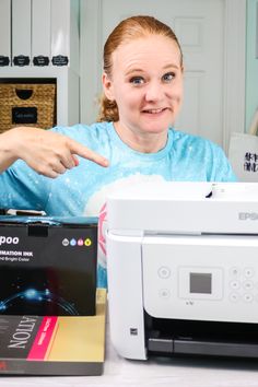 a woman pointing at a printer on the table next to it is another printer in front of her