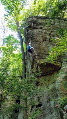 a man climbing up the side of a cliff with trees around him and another person standing next to him