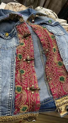 an old jean jacket with some gold and red beads on the bottom, sitting on top of a pile of clothes